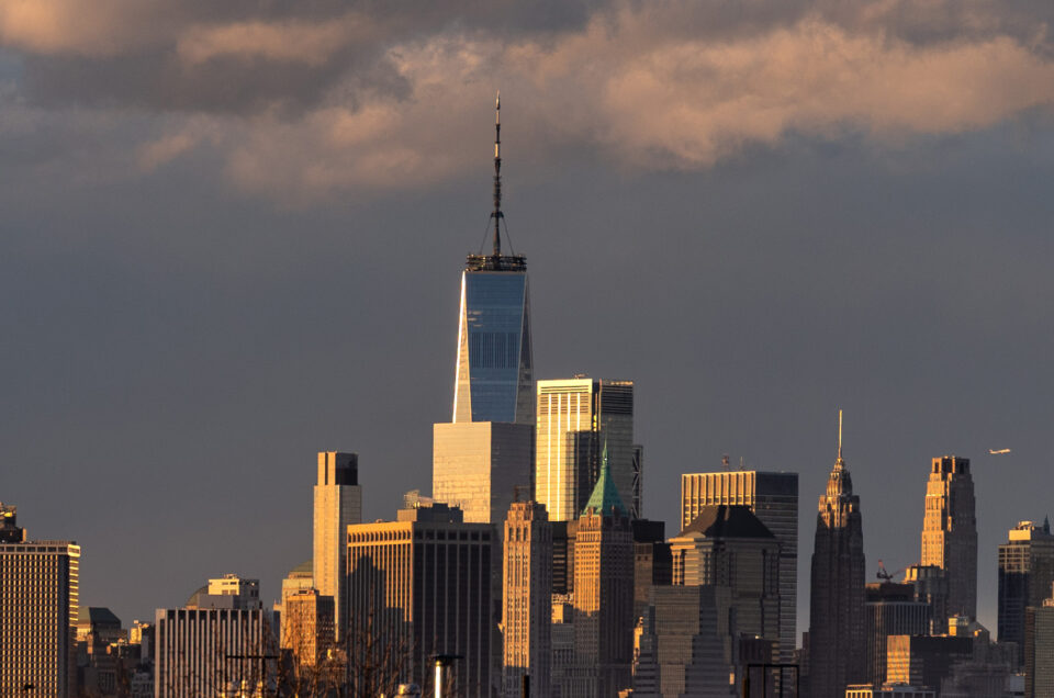 Manhattan skyline glowing gold at twilight, seen across the East River, with reflections on glass towers and clouds at sunset